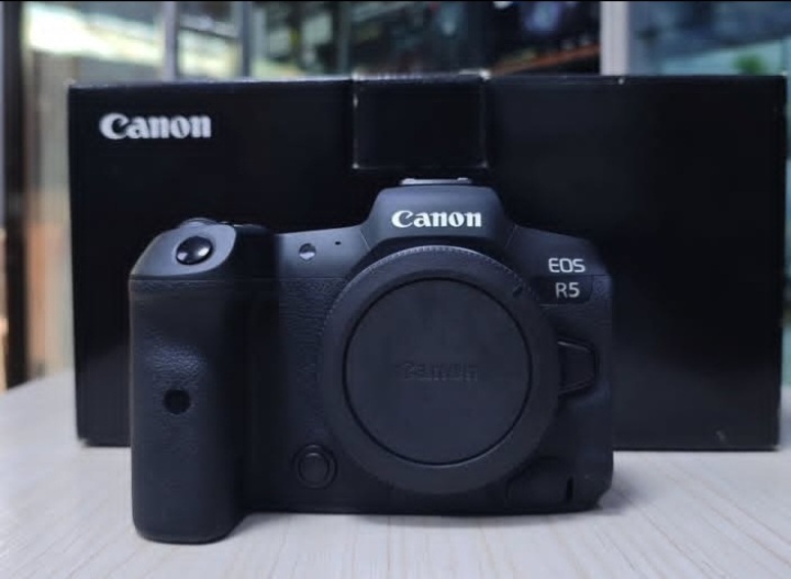 A black Canon EOS R5 mirrorless camera sits on a light wooden surface. Behind the camera is its original black box, which features the Canon logo printed in white.