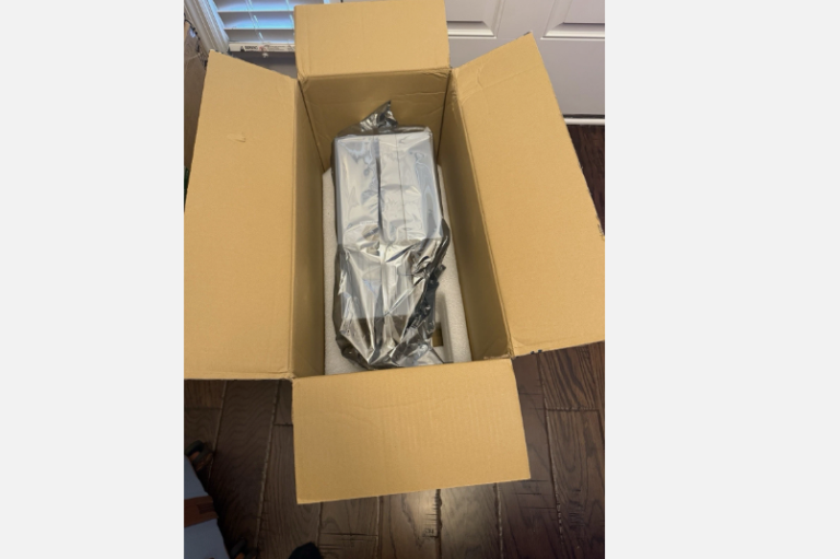 A rectangular object wrapped in a reflective silver bag sits inside an open cardboard shipping box. The item is cushioned by white foam padding and rests on a dark wood floor.